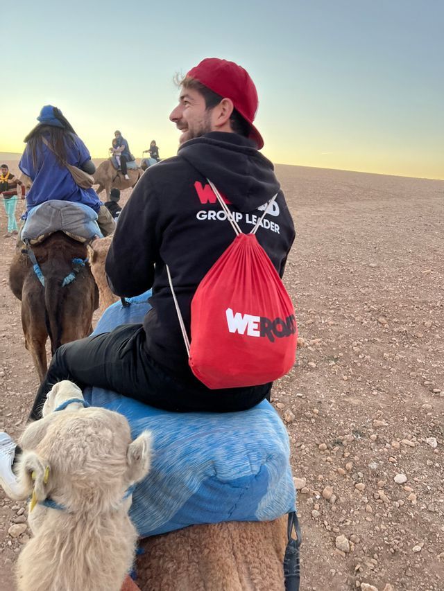 Un capogruppo WeRoad guida un viaggio di gruppo WeRoad a dorso di cammello attraverso un paesaggio desertico al tramonto.