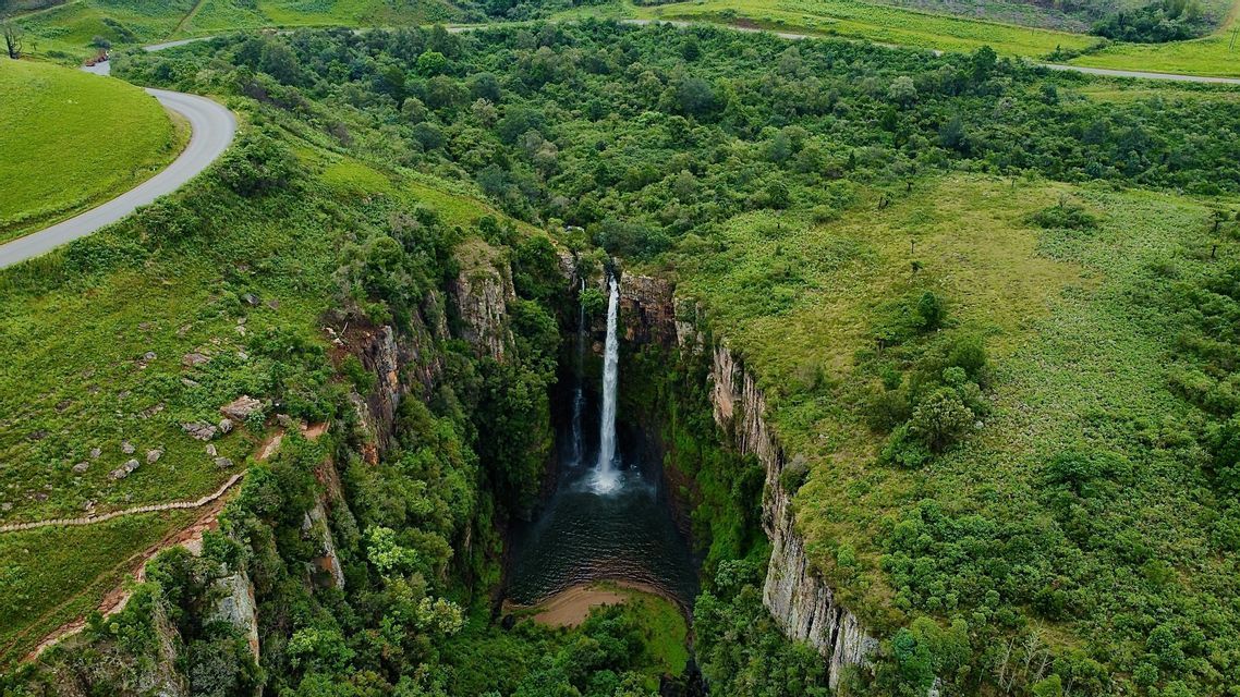 Luftaufnahme eines Wasserfalls, der von einer Klippe in ein Becken in einer üppig grünen Schlucht stürzt, umgeben von sanften Hügeln.