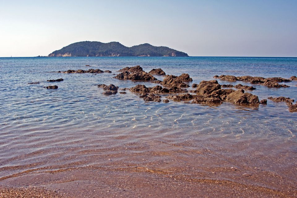 Una costa rocciosa con acqua limpida e poco profonda che lambisce una spiaggia sabbiosa, con un'isola verde all'orizzonte.