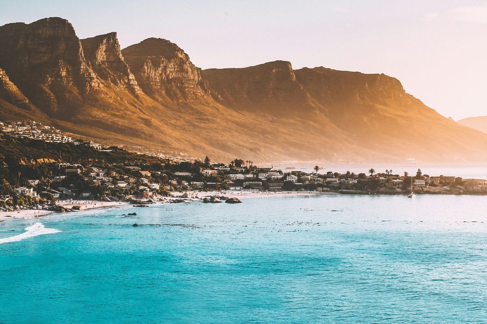Ein Panoramablick auf eine Küstenstadt mit einem weißen Sandstrand und türkisfarbenem Wasser am Fuße einer zerklüfteten Bergkette bei Sonnenaufgang.