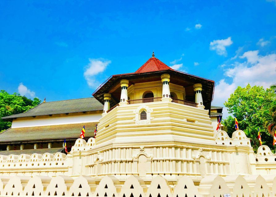 Un tempio bianco a più livelli con una torre ottagonale e tetto rosso si erge dietro un muro decorativo sotto un cielo blu.
