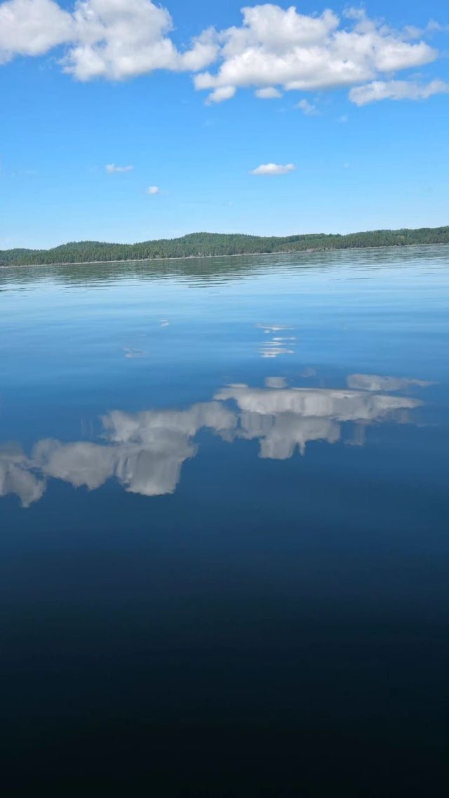 Ein ruhiger See spiegelt den blauen Himmel und die weißen Wolken darüber perfekt wider, mit einem bewaldeten Ufer im Hintergrund.