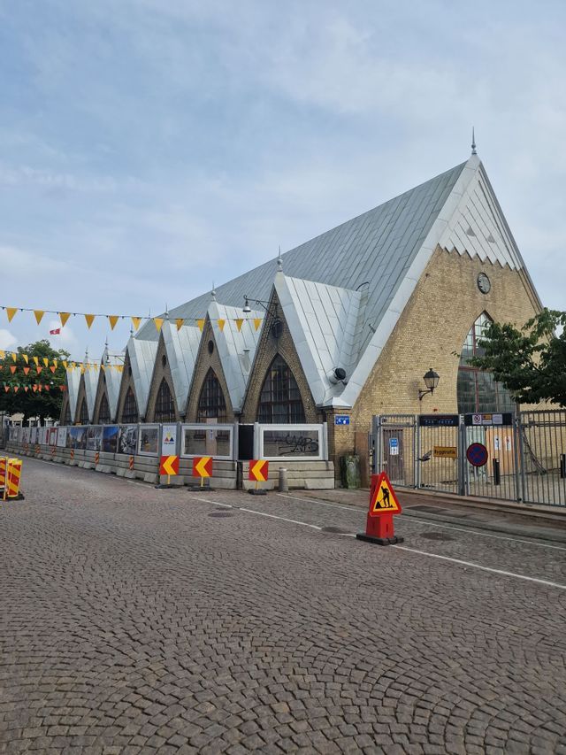 Ein großes Backsteingebäude mit mehreren Spitzgiebeln und einem Metalldach steht an einer Kopfsteinpflasterstraße, die von Bauzäunen gesäumt ist.