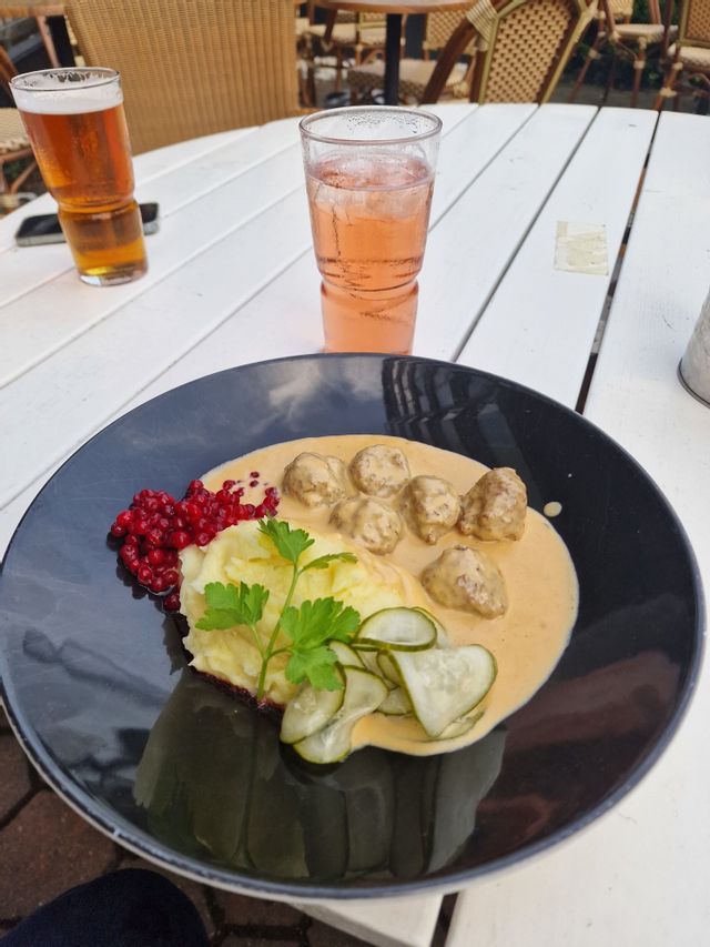 Ein Teller schwedische Köttbullar mit Kartoffelpüree, Preiselbeeren und Gurkenscheiben auf einem weißen Holztisch, dazu Getränke.