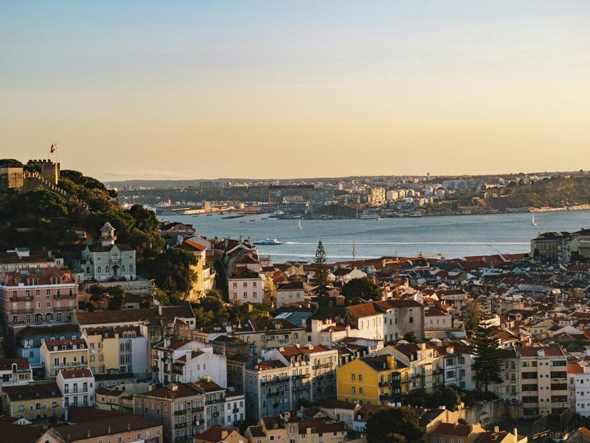 A cityscape of colorful buildings and red-tiled roofs overlooks a wide river at sunset, with a historic castle on a hill.