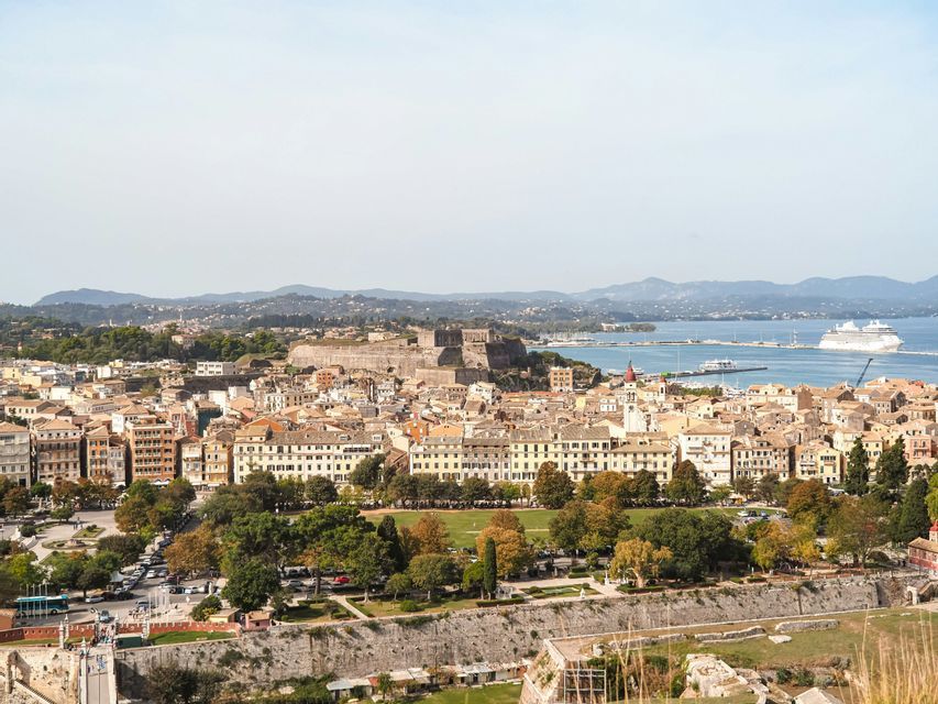An aerial view of a historic coastal city featuring a fortress, a park, and a large cruise ship in the harbor.