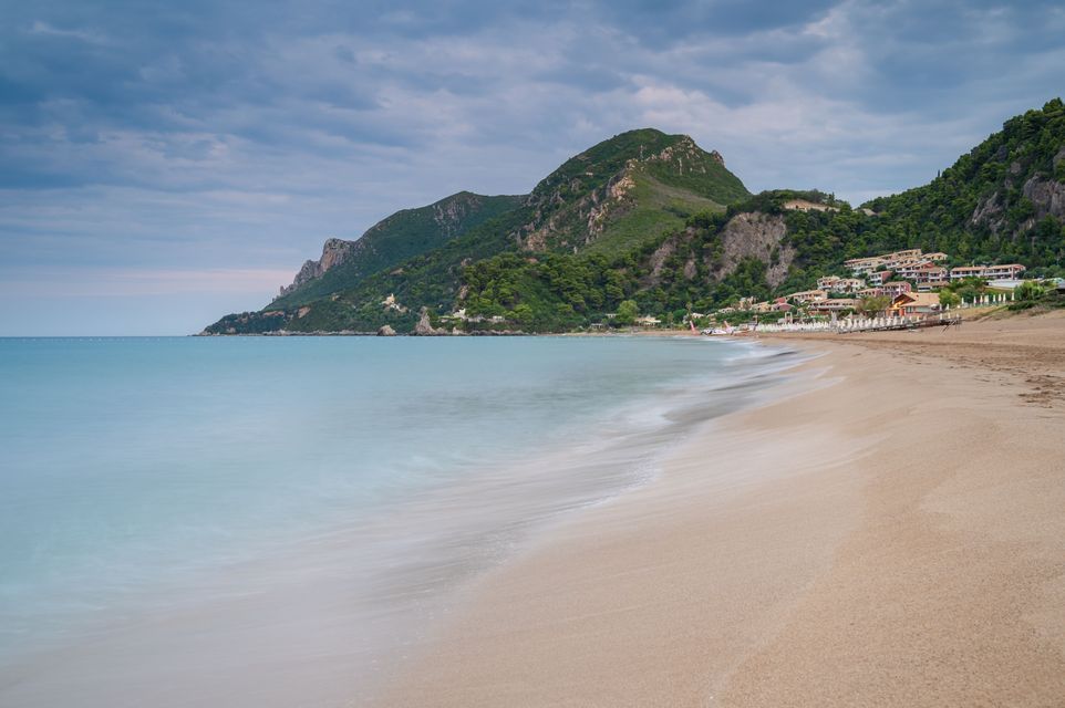 A long-exposure shot of turquoise water washing onto a sandy beach, with a green mountain and coastal buildings in the background.