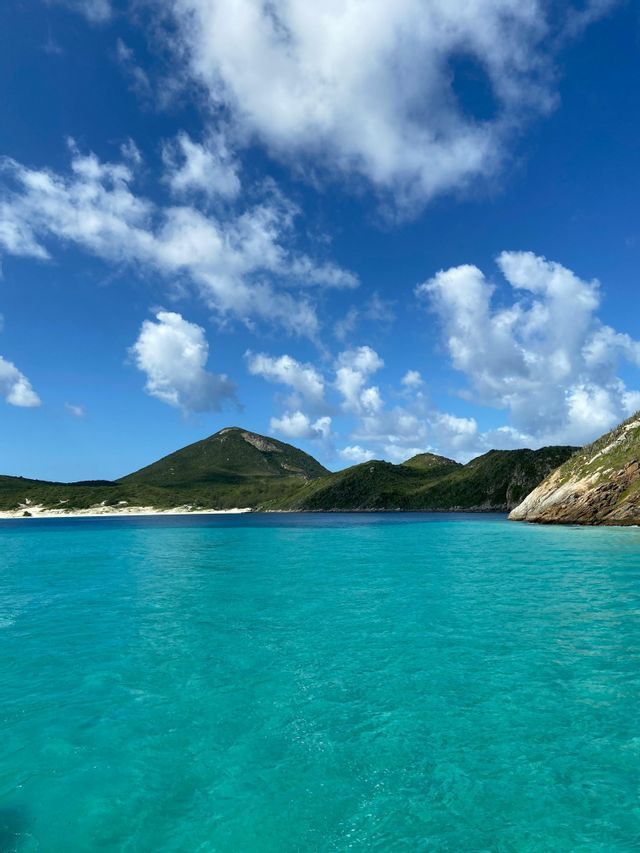 Agua turquesa cristalina en una cala, con colinas verdes y una playa de arena blanca al fondo bajo un cielo azul con nubes.