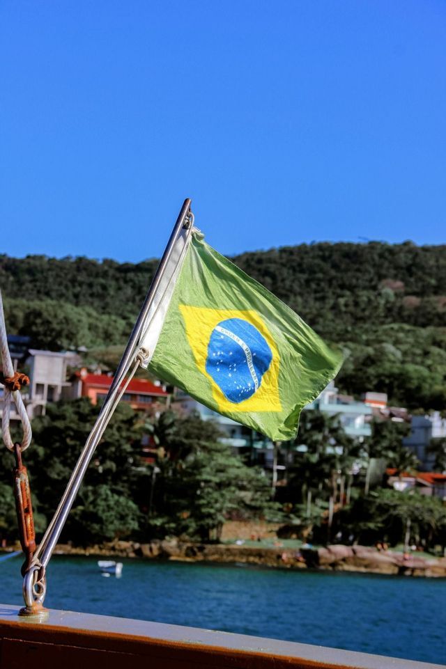 Una bandera de Brasil ondea en un barco, con el telón de fondo de una costa boscosa y un cielo azul.