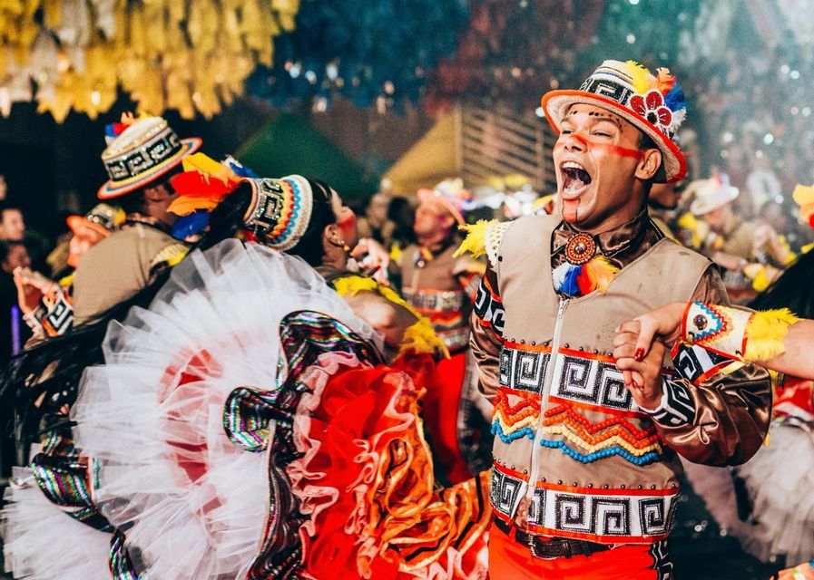 Un hombre con un colorido traje de plumas canta en voz alta mientras baila con una mujer durante un vibrante festival callejero.