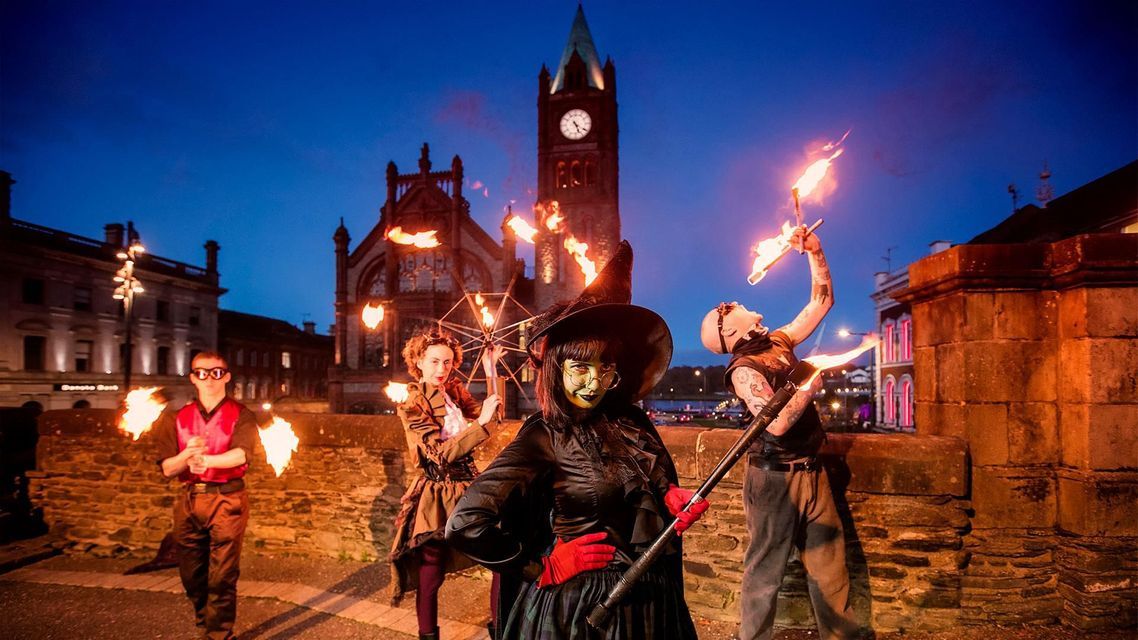 Quattro artisti in costumi elaborati manipolano torce infuocate e oggetti di scena di notte in una piazza cittadina con una torre dell'orologio.