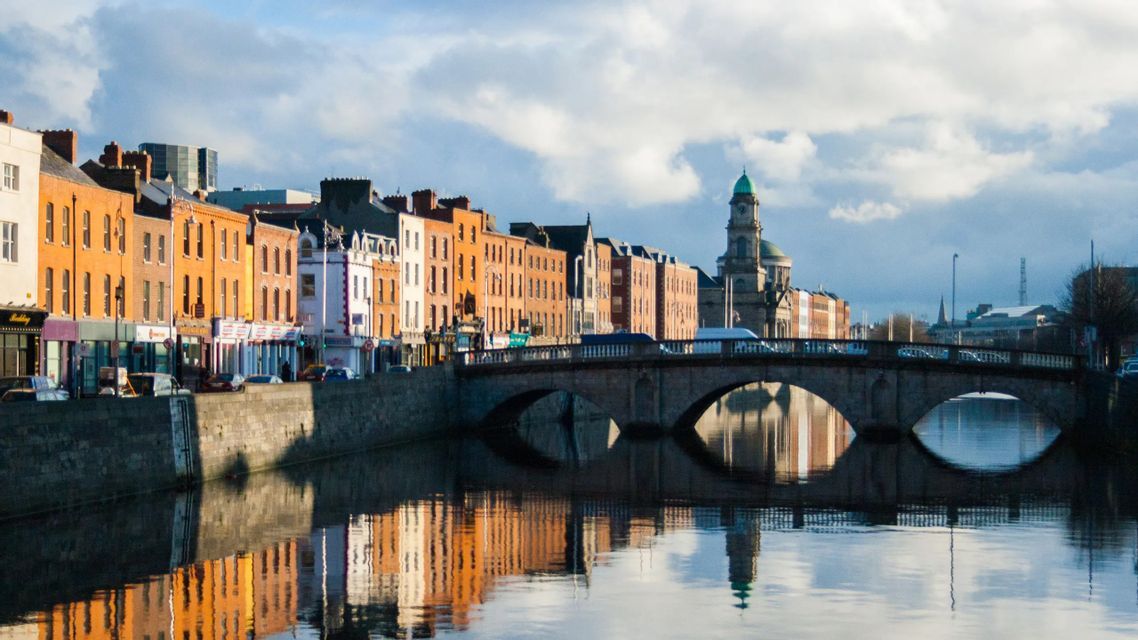 Una fila di edifici colorati e un ponte in pietra ad arco si riflettono nell'acqua calma di un fiume.
