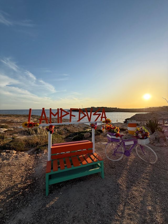 Un grande cartello con lettere arancioni, una panchina colorata e una bicicletta decorativa con fiori su una riva rocciosa al tramonto.