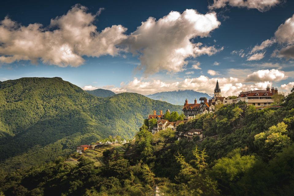 A collection of European-style buildings sits on a forested hillside overlooking a mountain range under a cloudy sky.
