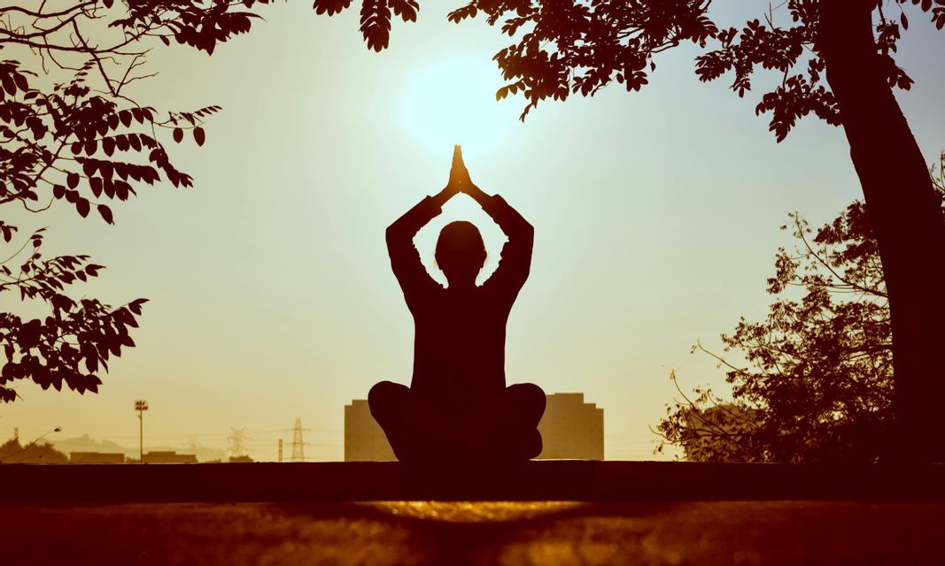 Silhouette di una persona in posa yoga all'alba, incorniciata da alberi con lo skyline della città sullo sfondo.