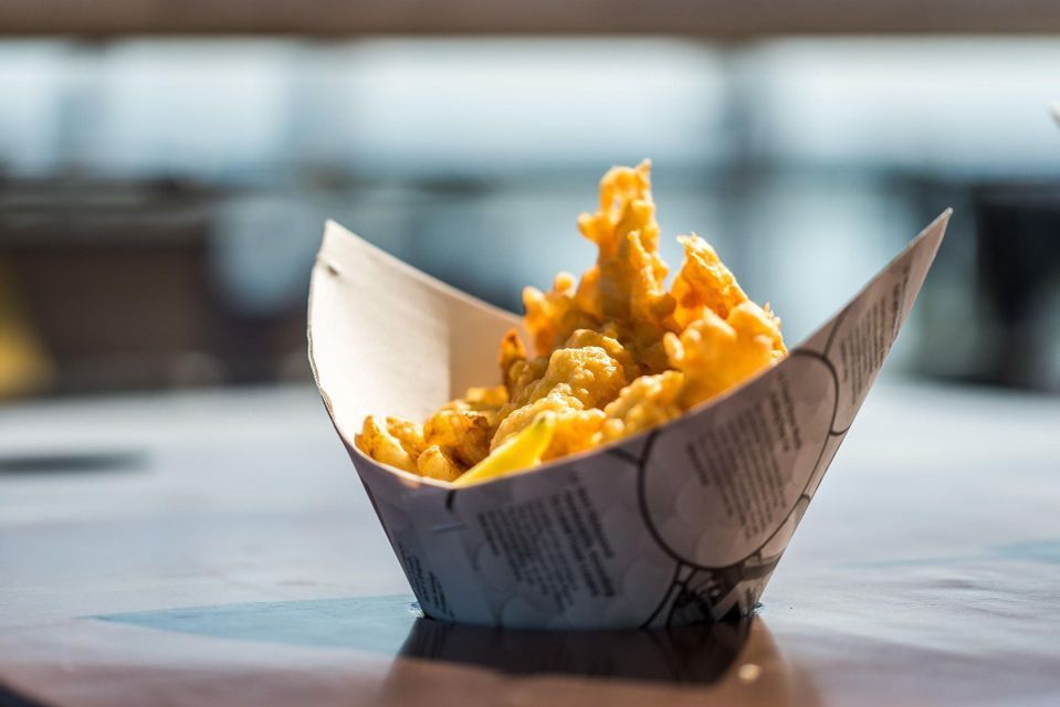 Un primer plano de comida frita dorada servida en un recipiente de papel estampado sobre una mesa oscura y reflectante.