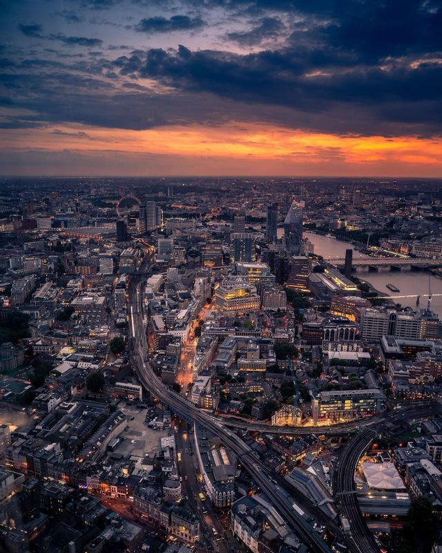 Una vista aérea de una extensa ciudad con calles y edificios iluminados bajo un dramático cielo al atardecer.