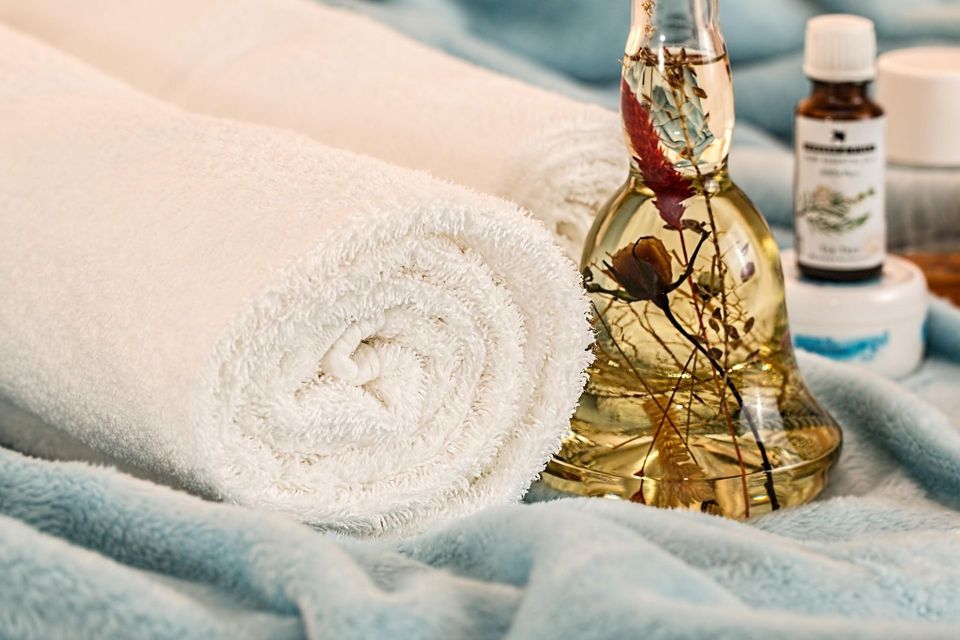 Primo piano di un asciugamano bianco arrotolato accanto a una bottiglia di olio da massaggio botanico e altri prodotti spa su una morbida coperta blu.