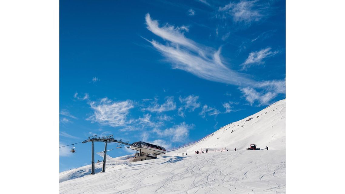 Una seggiovia e un gatto delle nevi su un pendio montano innevato, con sciatori sotto un cielo azzurro brillante e nuvole leggere.