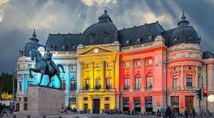 Ein Reiterdenkmal steht vor einem großen, prunkvollen Gebäude, das unter einem bewölkten Himmel in blauem, gelbem und rotem Licht erstrahlt.
