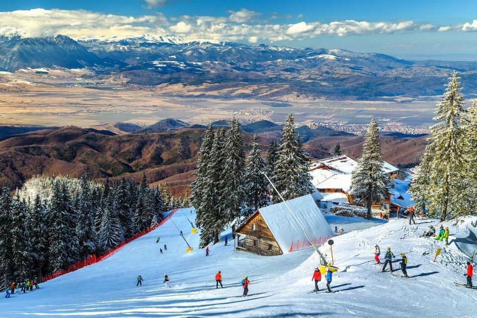 Skifahrer auf einem verschneiten Berghang mit Tannen und Chalets mit Blick auf ein weites Tal und ferne Gipfel unter blauen Himmel