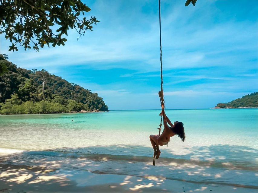 Une femme en bikini sur une balançoire en corde au-dessus d'une plage tropicale avec sable blanc et eau turquoise claire.