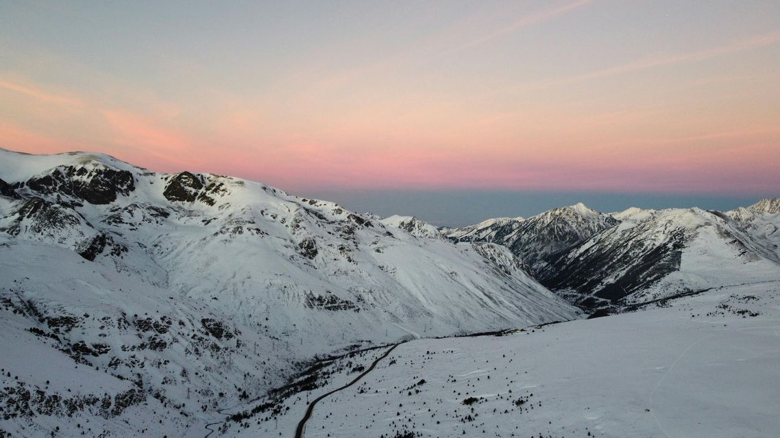 Una cordillera nevada con una carretera serpenteante en el valle bajo un cielo de atardecer rosa y morado.