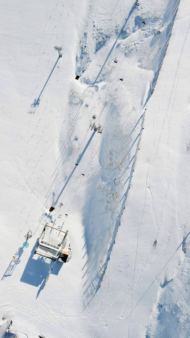Una vista aérea cenital de un telesilla y su estación sobre un vasto paisaje nevado proyectando largas sombras.