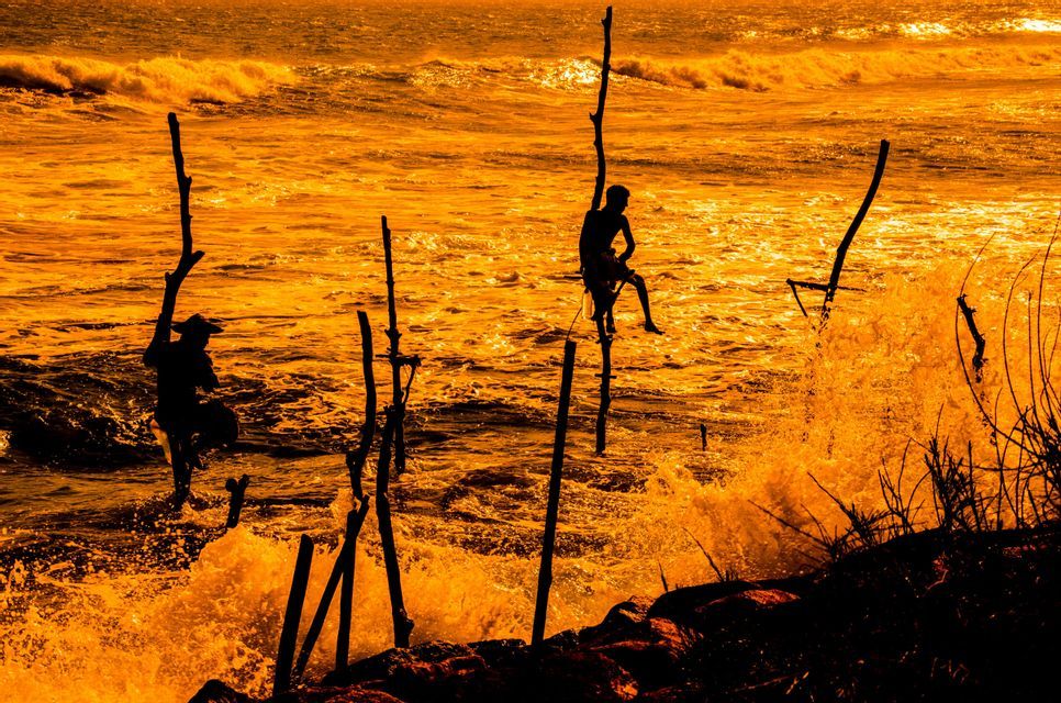 Sagome di due pescatori su palafitte in un mare mosso, arancione dorato, al tramonto, con le onde che si infrangono.