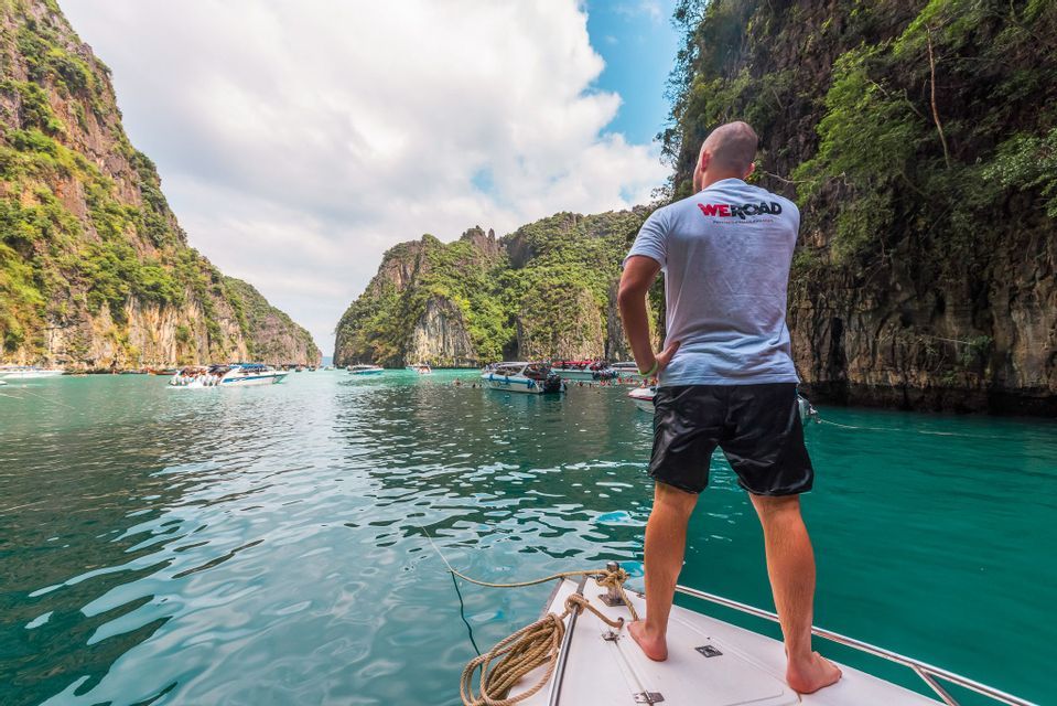 Un homme d'un voyage WeRoad se tient à la proue d'un bateau, contemplant une baie turquoise aux falaises rocheuses.