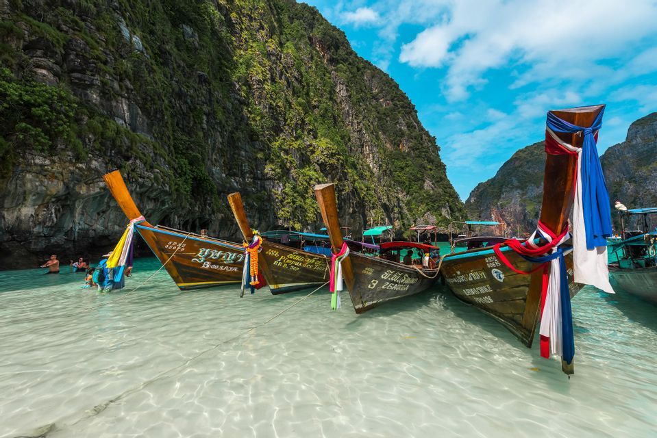 Eine WeRoad-Gruppenreise schwimmt in einer klaren Lagune neben traditionellen Longtail-Booten, die an steilen grünen Klippen festgemacht sind.