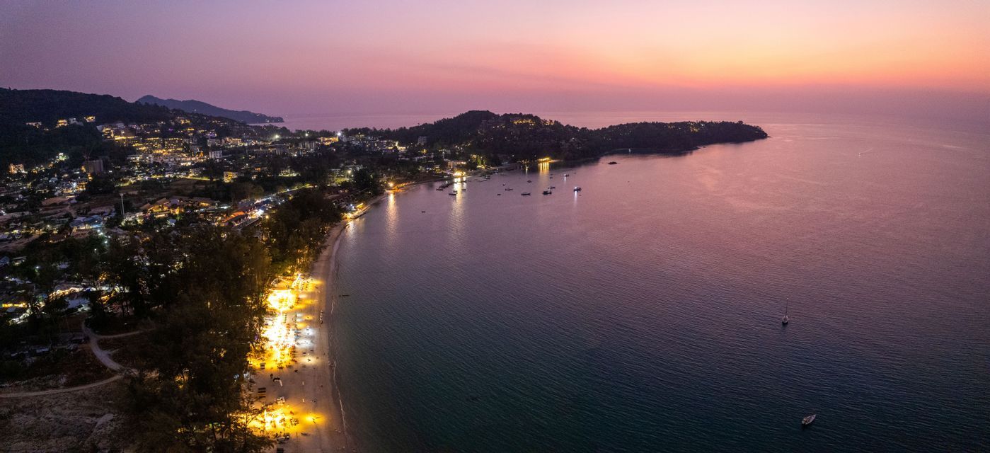 Vue aérienne d'une ville côtière lumineuse et de sa plage, bordant une baie calme avec des bateaux au crépuscule.