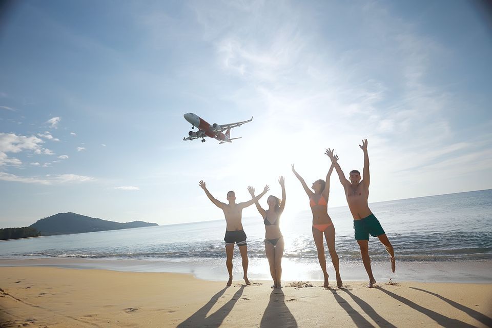 A WeRoad group trip waving with arms raised on a beach as a large airplane flies low overhead.