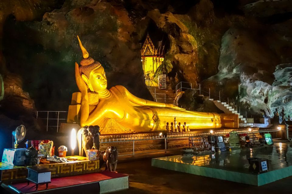 A large, golden reclining Buddha statue illuminated by artificial lights inside a dark, rocky cave.