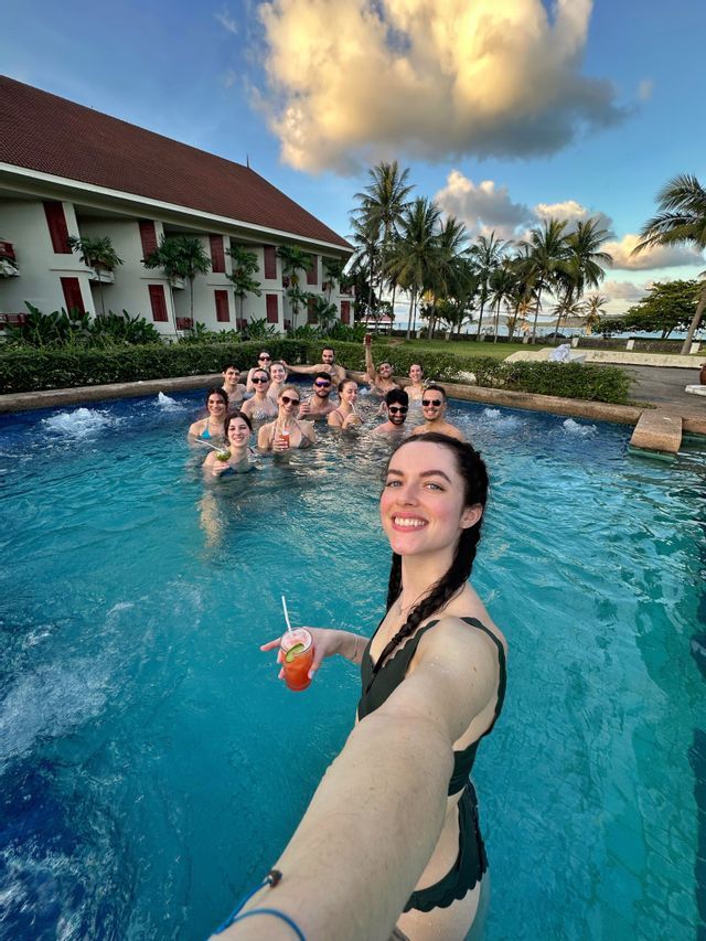 A woman holding a cocktail takes a selfie with a WeRoad group trip in a resort swimming pool.
