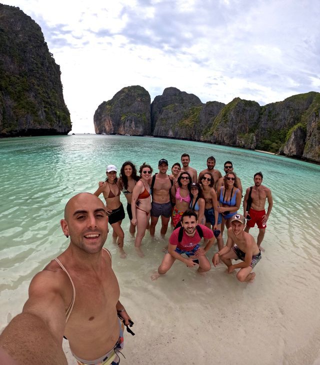 A man takes a selfie of his WeRoad group trip standing in the shallow, clear water of a tropical bay surrounded by cliffs.