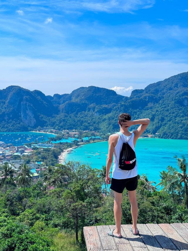 Ein Mann mit Rucksack steht auf einem Aussichtspunkt und blickt über eine tropische Landenge mit zwei türkisfarbenen Buchten und grünen Bergen.