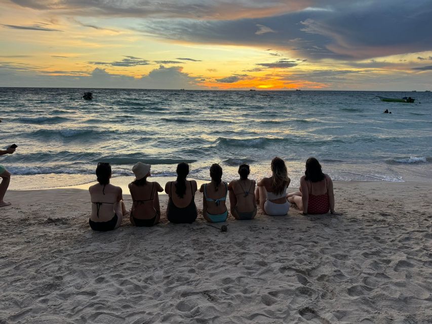 A WeRoad group trip of women sitting on a beach, facing the ocean and watching the sunset.