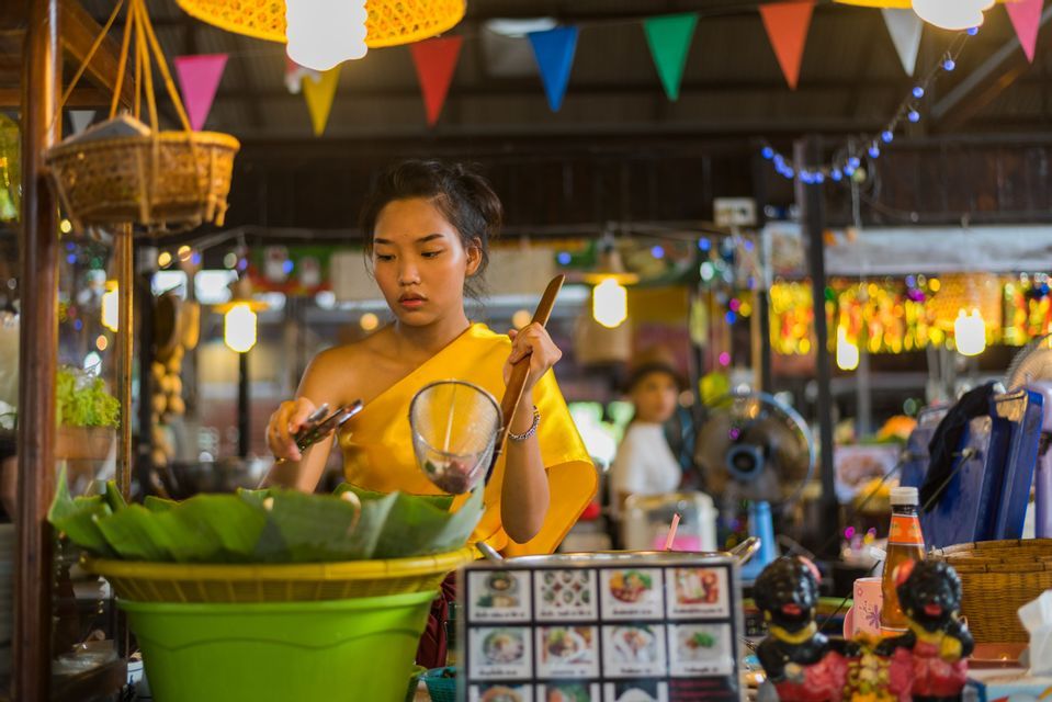 A woman in a yellow top prepares food at an indoor market stall decorated with colorful lights and banners.