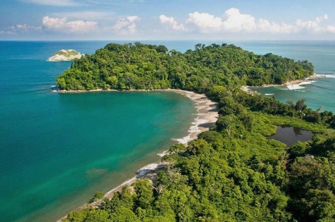 An aerial view of a tropical peninsula covered in dense forest, featuring a curved sandy beach and turquoise ocean water.