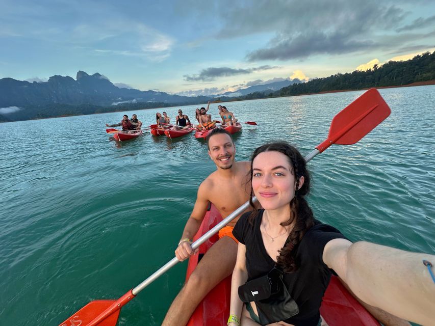 A WeRoad group trip takes a selfie while kayaking in red boats on a lake, with mountains in the background under a cloudy sky.
