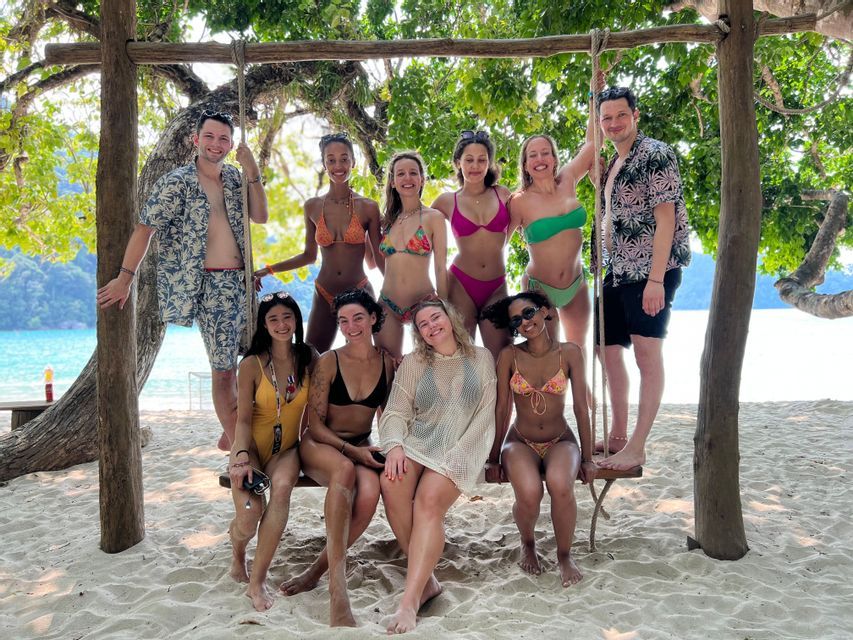 A WeRoad group trip of ten people in swimwear posing together on a large wooden swing on a sandy beach.