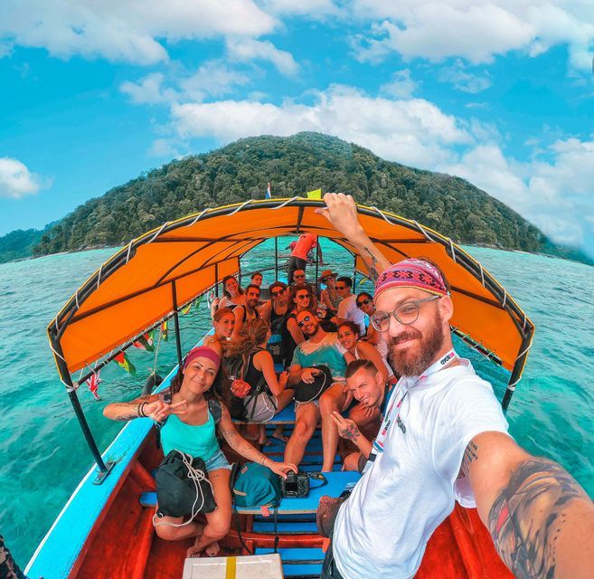 A WeRoad group trip smiles for a selfie on a boat with an orange canopy, cruising on turquoise water by a forested island.