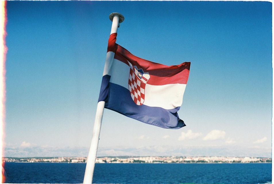 Die kroatische Flagge weht an einem Fahnenmast vor klarem blauem Himmel, mit dem Meer und einer fernen Küstenlinie im Hintergrund.