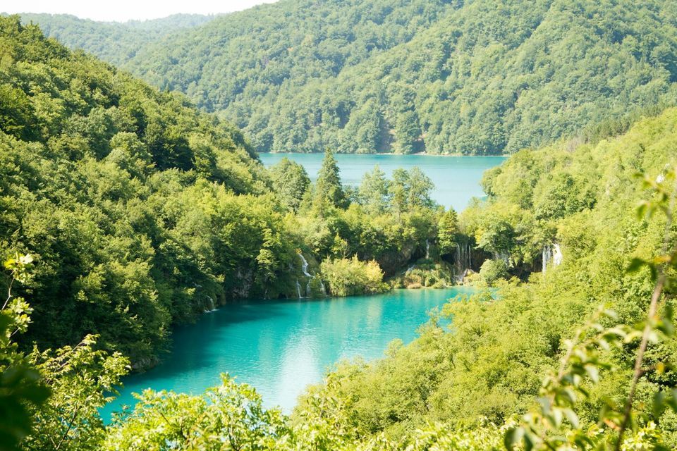 Ein Blick von oben auf einen türkisfarbenen See, umgeben von dichten grünen Wäldern, in den kleine Wasserfälle münden.