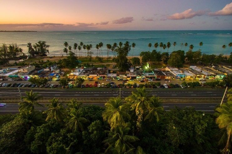 Una veduta aerea di una strada costiera e di una città con negozi illuminati, accanto a una spiaggia con palme al tramonto.