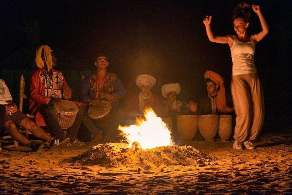 Un grupo de WeRoad se reúne de noche, donde una mujer baila junto a una hoguera mientras unos hombres tocan tambores en la arena.