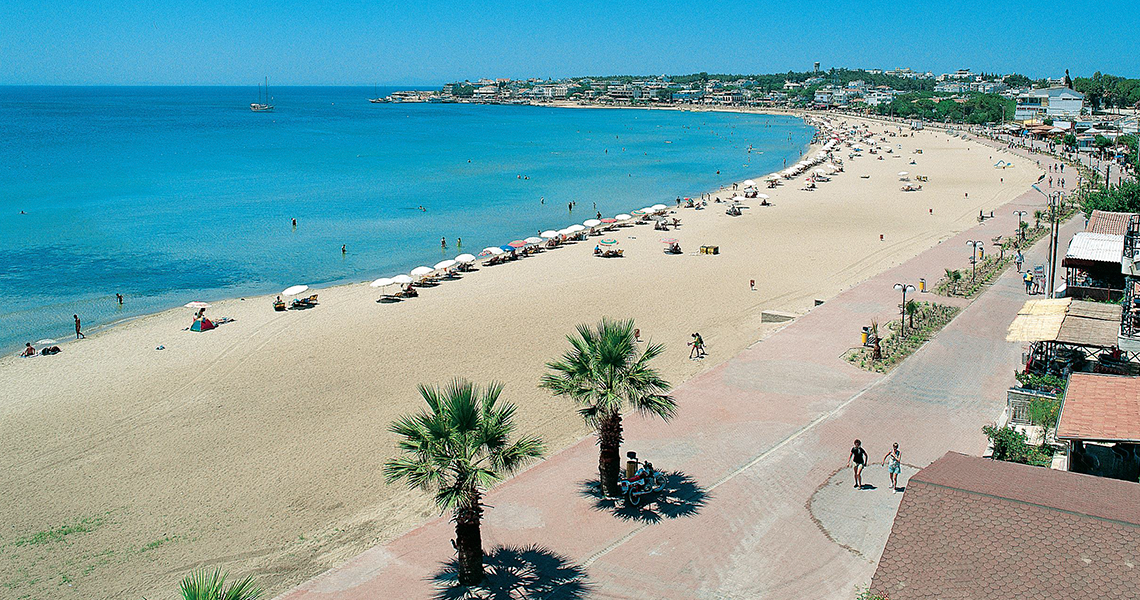Vue aérienne d'une longue plage de sable incurvée avec des gens sous des parasols, à côté d'une mer turquoise calme et d'une promenade bordée de palmiers.