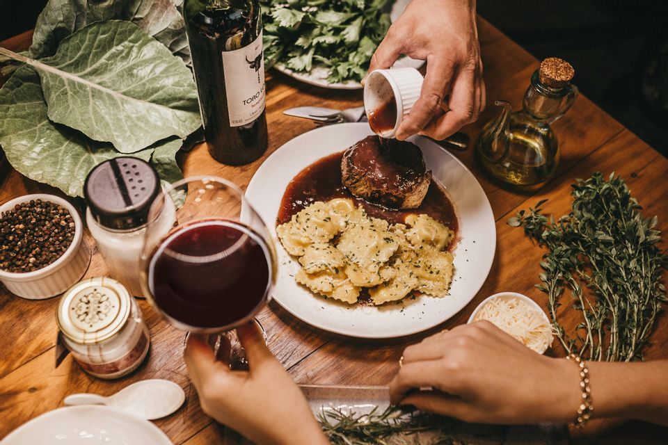 Una persona versa la salsa su un piatto di filetto mignon e ravioli su un tavolo di legno apparecchiato per cena con vino rosso.
