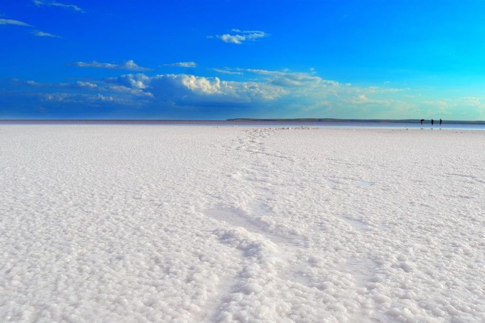 Des empreintes serpentent sur une étendue de sel blanc texturée vers trois silhouettes lointaines à l'horizon sous un ciel bleu éclatant.